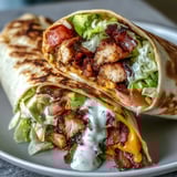 Golden-brown Crispy Chicken Bacon Wrap sliced in half, revealing creamy avocado slices and juicy diced tomato on a warm, fluffy tortilla.