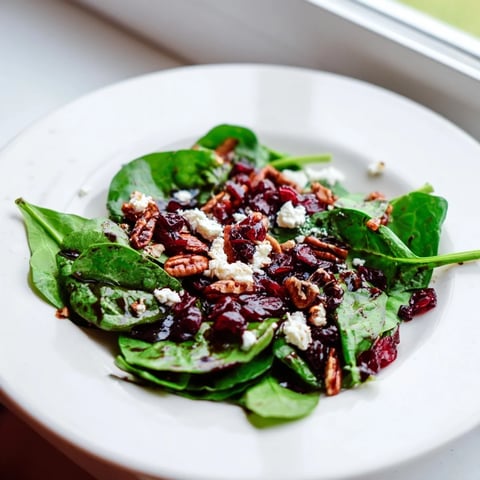 Fresh baby spinach, creamy goat cheese, cranberries and candied pecans tossed in tangy balsamic vinaigrette, in a white ceramic salad bowl.